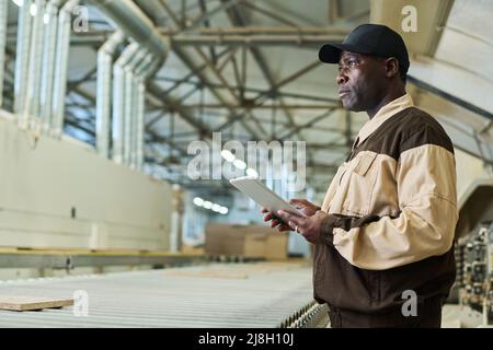 Serious African construction worker in uniform working on digital tablet while standing in workshop of factory Stock Photo
