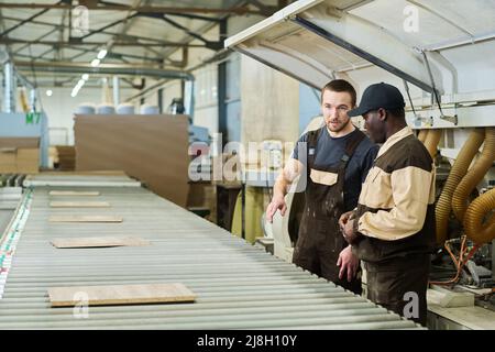 Worker in unifrm discussing production of details with foreman while they standing next to machine at factory Stock Photo