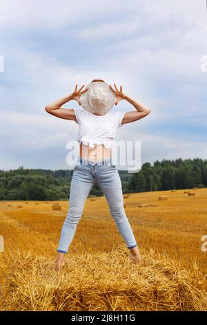 barefoot girl in straw hat stands on a haystack on a bale in the agricultural field after harvesting. Stock Photo