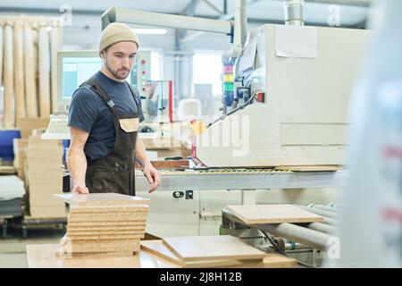 Young man in overall working with timber at factory standing at machine Stock Photo