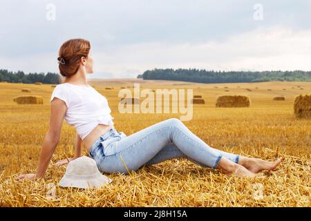 barefoot girl with straw hat sitting on a haystack on a bale in the agricultural field after harvesting. Stock Photo