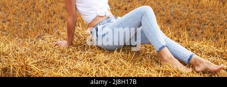 barefoot girl sitting on a haystack on a bale in the agricultural field after harvesting. banner Stock Photo