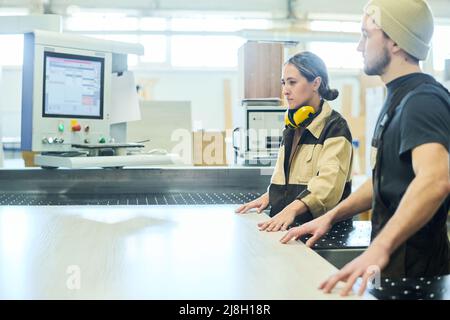 Two young workers in uniform working in team with wooden board on cutting machine at furniture factory Stock Photo