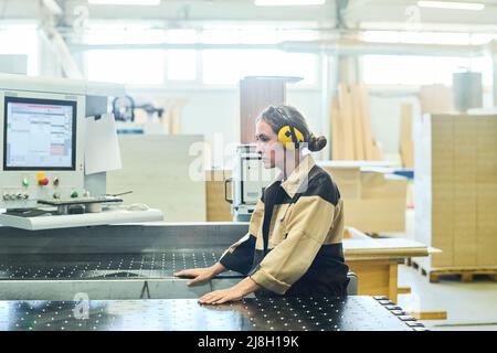 Young female worker in protective headphones standing at machine in the plant Stock Photo