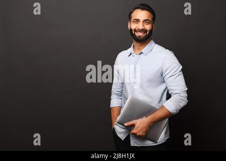 Cheerful Indian bearded man holding laptop standing isolated on black, looking at camera with happy smile. Male employee or freelancer carrying laptop computer. Copy space Stock Photo