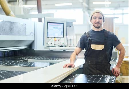 Portrait of a young carpenter in an apron, smiling at the camera, standing at the modern furniture production equipment Stock Photo