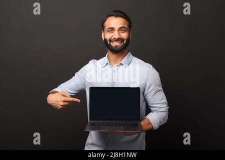 Happy Indian man holding laptop with empty screen and points finger at it, hispanic male employee presenting new computer app, promotion and advertise concept Stock Photo