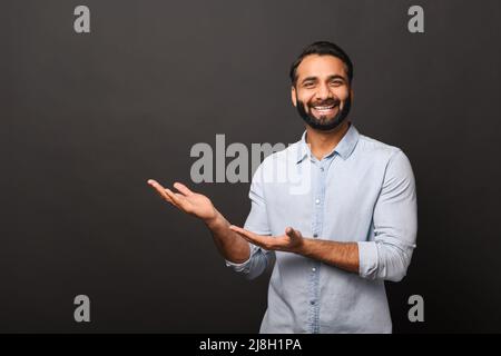 Happy Indian bearded man in casual shirt points hands aside at empty space, hispanic man advertising novelty standing isolated on black, showing with palms sideway Stock Photo