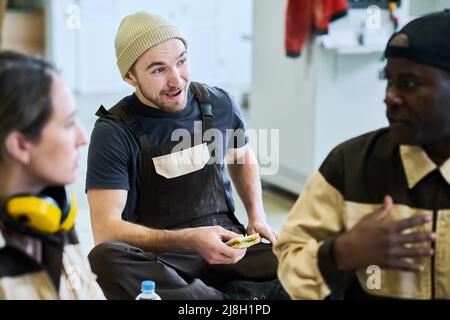 Young man in overall eating sandwich and talking to his colleagues during lunch at warehouse Stock Photo