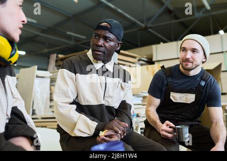 African mature worker in uniform having lunch with his colleagues at warehouse, they resting after work Stock Photo