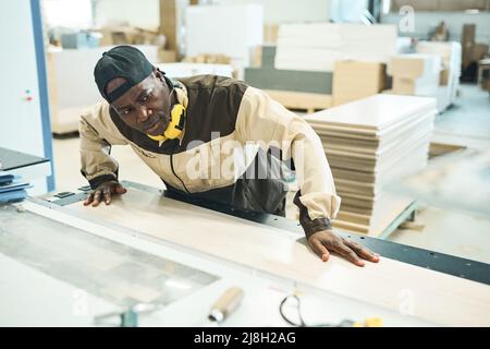 Serious African man in uniform controlling the process of sawing the wooden plank on machine at factory Stock Photo