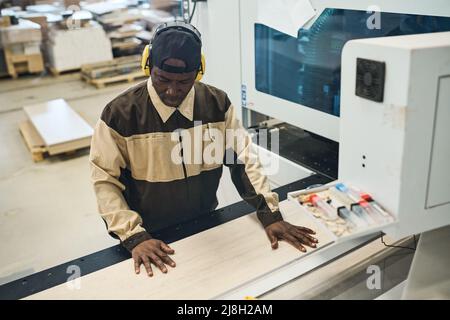 African craftsman in protective headphones and uniform concentrating on his wood work at machine Stock Photo