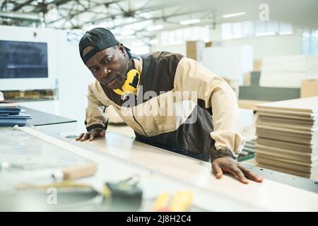 Serious African carpenter sawing wood on special machine during his work at furniture factory Stock Photo