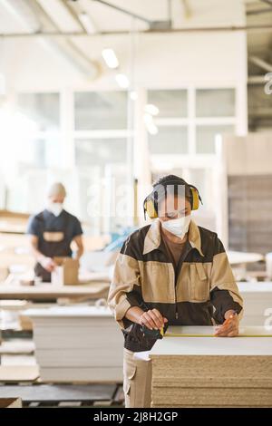 Young female worker in mask and headset measuring wooden plank before sawing during her work at factory Stock Photo