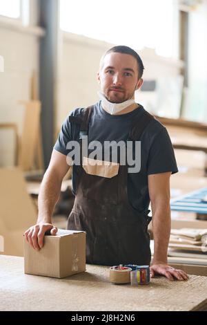 Portrait of young worker in overall looking at camera while packing box at table in warehouse Stock Photo