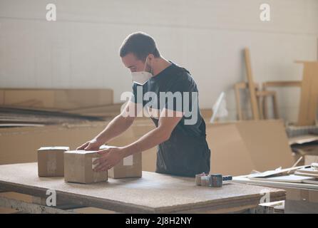 Young man in protective mask preparing packages for shipping, he working at table in warehouse Stock Photo