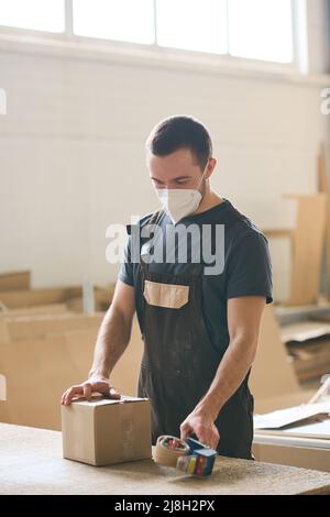 Young man in uniform standing at table and packing box with adhesive tape before shipping Stock Photo