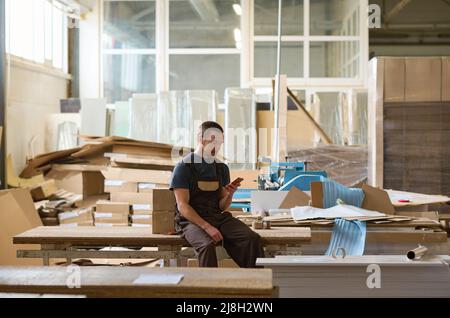Manual worker sitting on table and using his mobile phone during his break after work in warehouse Stock Photo