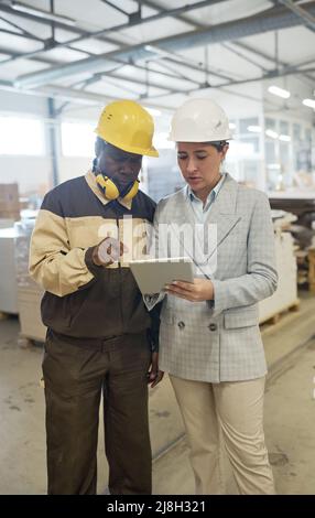 Young engineer in helmet pointing at digital tablet and discussing project with worker while they standing at workshop Stock Photo