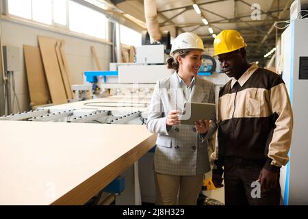 Engineer controlling the work at factory using tablet pc, she talking to foreman while they standing at machine Stock Photo