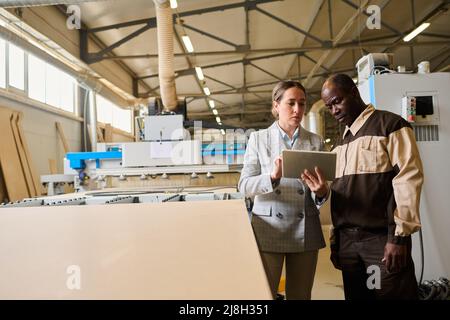 Engineer using digital tablet and discussing project together with foreman in uniform while they standing near the equipment at plant Stock Photo
