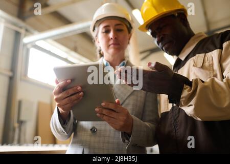 African foreman in hardhat pointing at tablet pc and talking to engineer during work at factory Stock Photo