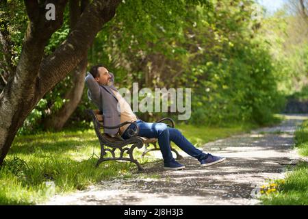 Handsome mature freelancer man with grey beard, sitting on the bench at the park with hands folded behind his head enjoying free time wearing casual jacket and jeans.  Stock Photo