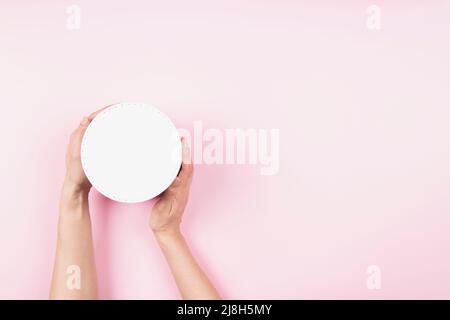 Woman hands holding white round cardboard box with cap onpastel pink background. Mock up package box. Top view Stock Photo