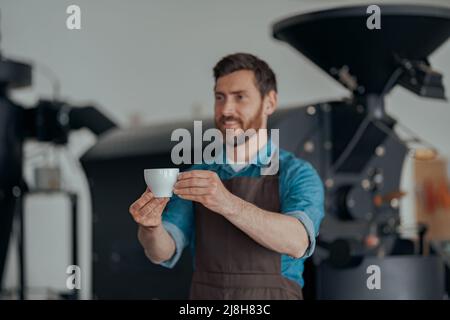 Barista propose cup of fresh coffee on the background of roasting coffee machine and looking away Stock Photo