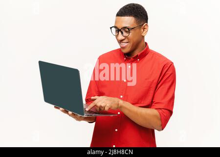 Cheerful handsome smiling African-American young man in red shirt using laptop isolated on white. Friendly smart multiracial student, freelancer in glasses typing, using computer for remote work Stock Photo