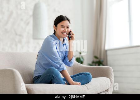 Positive korean lady sitting on sofa, talking on phone Stock Photo