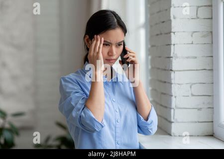 Upset asian woman standing by window, having phone conversation Stock Photo