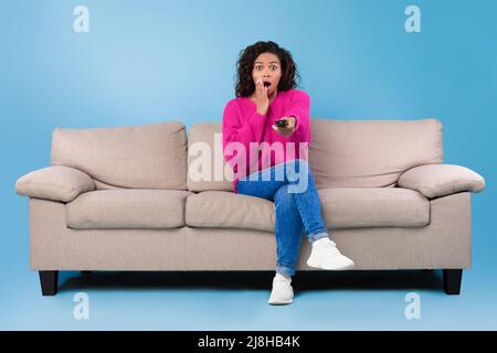 Frightened young African American lady with remote control sitting on sofa and watching scary movie or thriller on TV Stock Photo