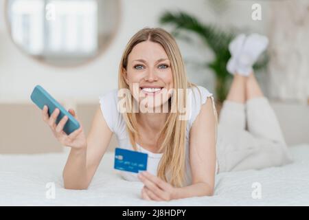 Satisfied pretty young blond caucasian woman with smartphone and credit card lying on bed in white bedroom Stock Photo