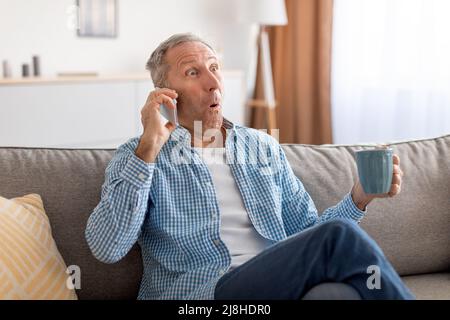Shocked mature man talking on cellphone sitting on sofa Stock Photo