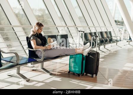 Passenger sitting on chair in modern light airport with outstretched legs on suitcases, reading magazine and waiting airplane arriving, flight delay Stock Photo