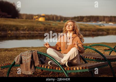 Beautiful blonde woman sitting alone on bench next to river bank on cool sunny day, relaxing, meditating and enjoying peace and freedom. Outdoors Stock Photo