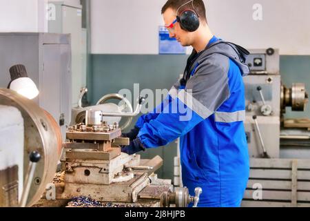 Young turner in safety glasses and headphones works at lathe in workshop. Authentic scene workflow. Caucasian worker processes metal parts. Stock Photo