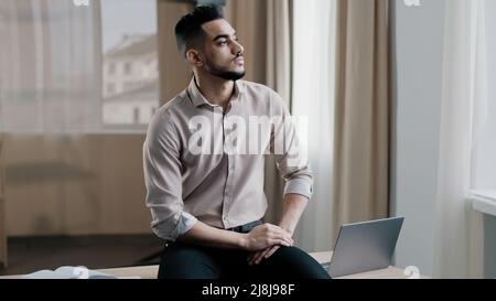 Thoughtful young man sad professional worker sitting on desk looking out window deep in thoughts recollect memory pondering problem solution Stock Photo