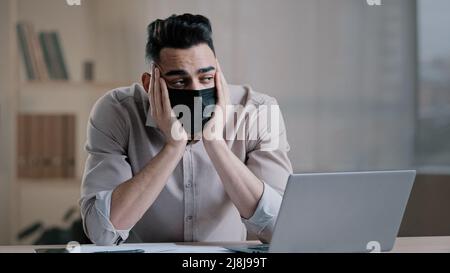 Bored tired hispanic guy worker in medical mask during pandemic coronavirus young business man tired ill worker feel boredom in home office hold head Stock Photo