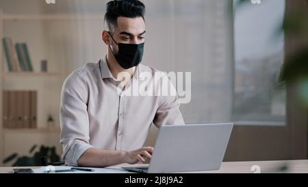 Hispanic arab male business man masked worker programmer typing on laptop search online information in cyberspace developing corporate project using Stock Photo