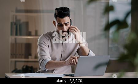 Busy millennial arabian business man web designer employee using laptop talking on cell phone involved in multitasking using gadgets discussing online Stock Photo