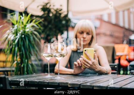 Adult mature woman texting messaging at mobile phone in bar outdoors with wine glasses and blurry restaurant, drinking white wine. People using Stock Photo