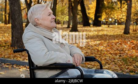 Happy outdoors elderly middle aged woman joyfully resting in autumn park sitting in wheelchair with disability illnesses single mature senior grey Stock Photo