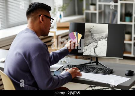 Back view at male editor comparing color swatches to pictures on computer screen while working in office, copy space Stock Photo