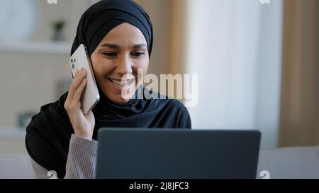 Happy smiling arabian woman muslim girl in hijab female freelancer talking by mobile phone with friend answering call enjoy good news has business Stock Photo