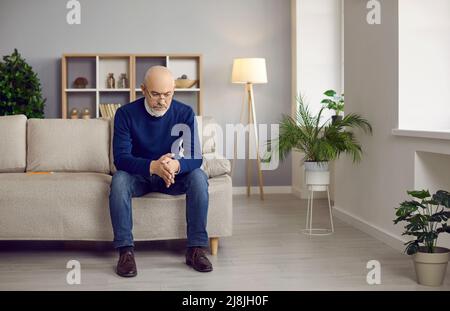 Older man with serious expression sits on sofa at home and thinks about his life and business. Stock Photo