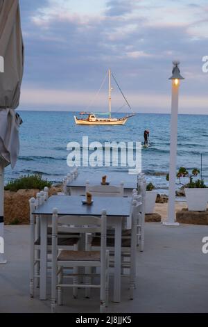 Seaside restaurant tables paddle surfer and a sailboat bathed in afternoon light in Potamos Liopetri fishing village in the Mediterranean island of Cy Stock Photo