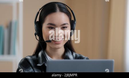 Portrait asian woman student girl online distant teacher wear headset talking conference call speak looking at laptop at home office video chat job Stock Photo