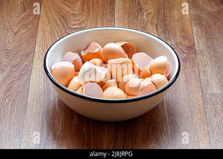 a white tureen with eggshells stands on a wooden kitchen countertop of a brown table, the remains of used chicken eggs are broken cracked shells after Stock Photo